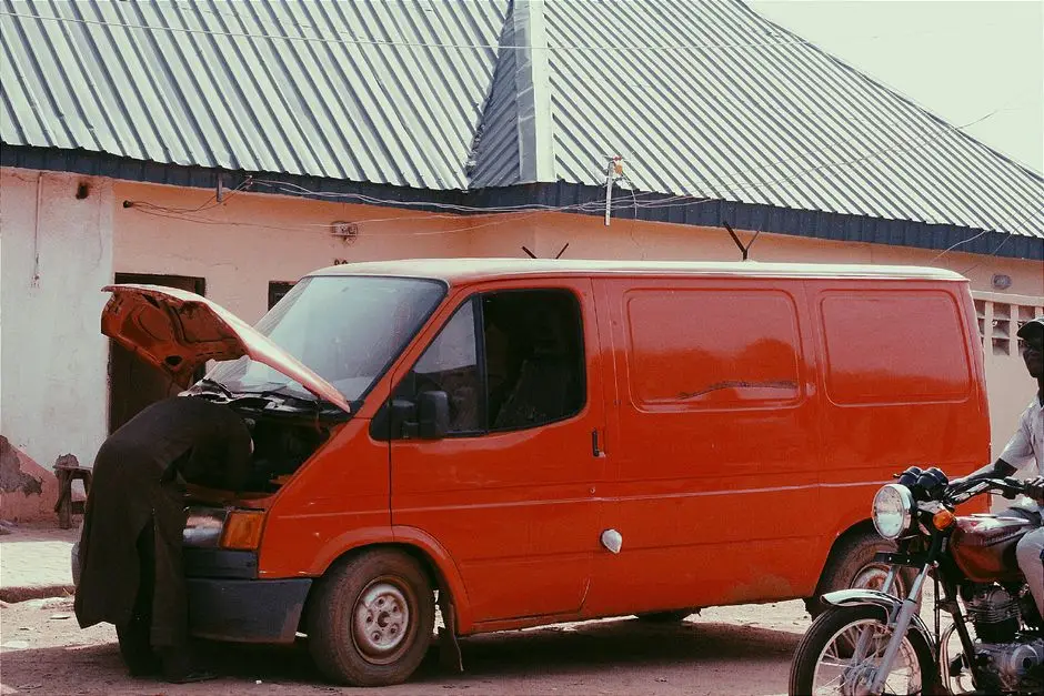 Mechanic repairing a red van on a city street with a motorbike nearby.